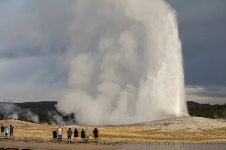 Old Faithful erupting under a stormy sky as visitors watch from the boardwalk
