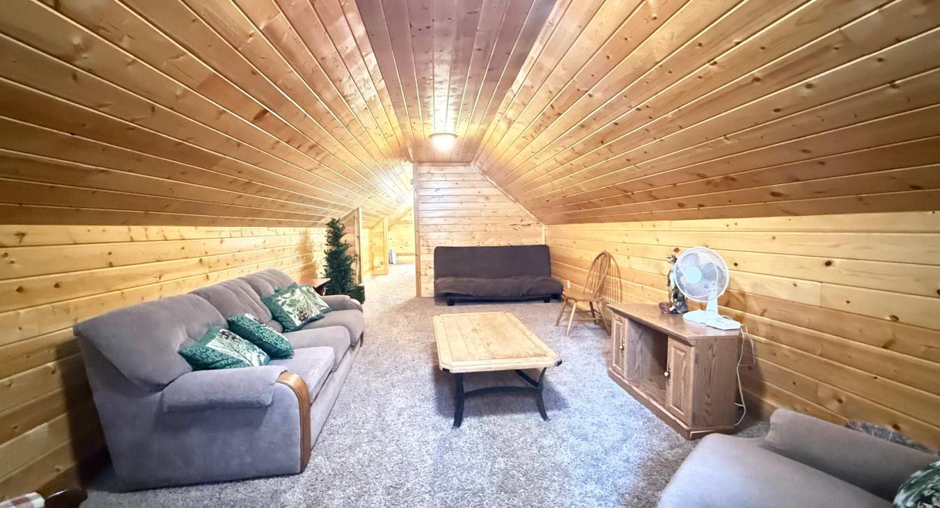 This comfortable garage loft lounge features a cozy sleeper sofa, plush seating, and a TV setup, nestled under beautiful sloped wood ceilings.