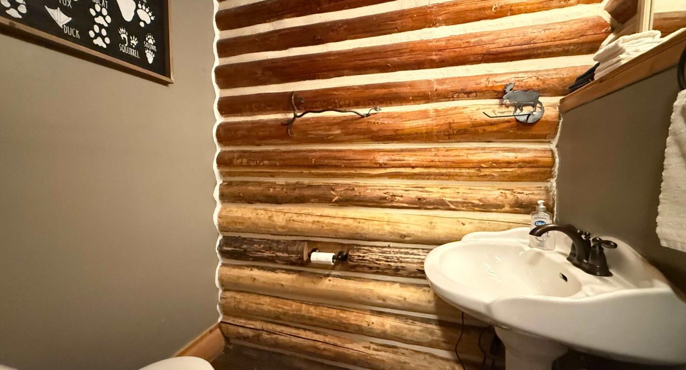 The convenient half bathroom highlights authentic log-cabin craftsmanship, complete with a classic pedestal sink, toilet, and charming forest-themed decor.
