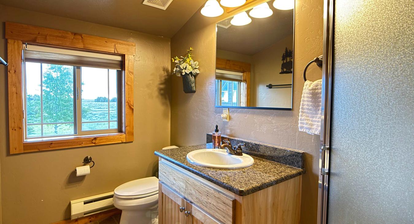 The convenient garage studio bathroom features a beautiful granite-top vanity, large mirror, toilet, and plenty of warm lighting.