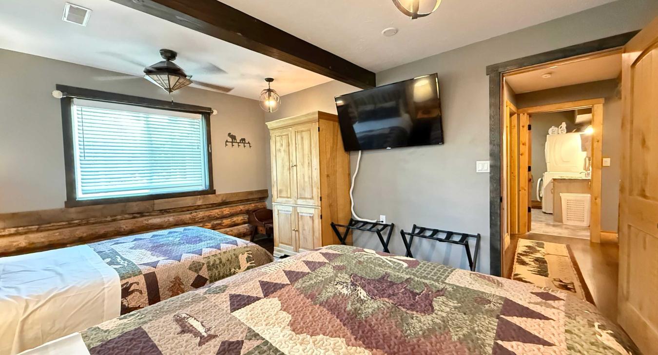 The dual-Queen room features a classic wooden armoire for storage, a wall-mounted TV, and easy access to the nearby laundry hallway.
