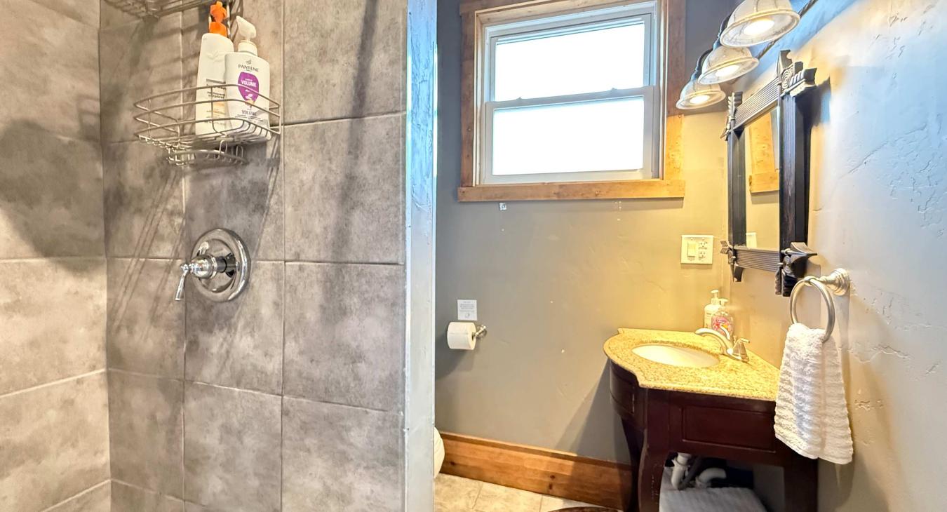 The pristine 3/4 bathroom features a tiled walk-in shower, a beautiful dark wood vanity with a granite top, and bright vanity lighting.